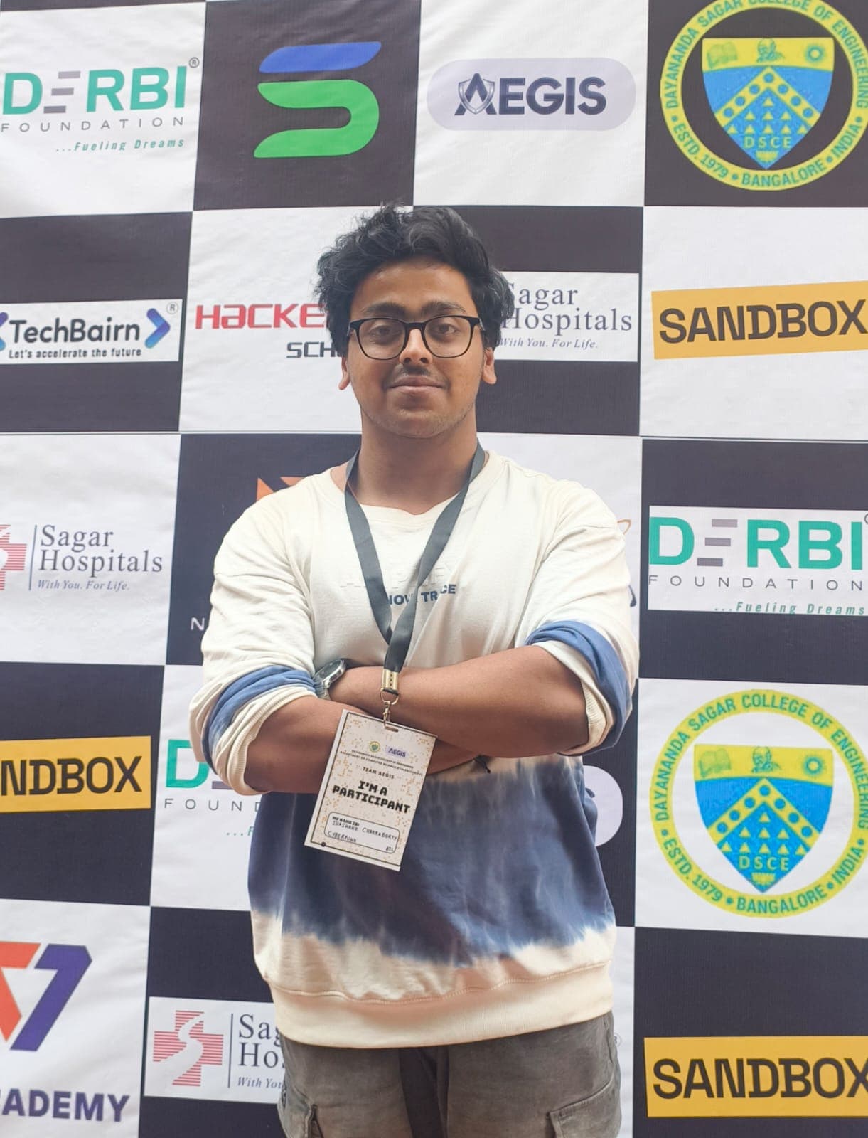 Shashank standing arms-crossed in front of the Sandbox sponsor backdrop, participant badge around his neck.